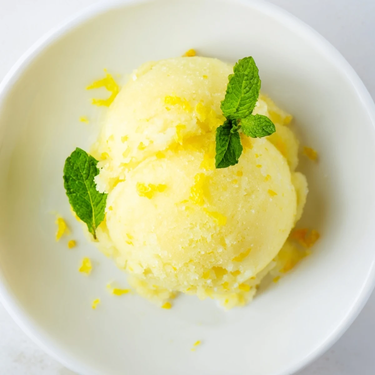 Bright yellow lemonade sorbet in a clear glass bowl, topped with fresh mint leaves and a lemon twist for a refreshing summer treat.