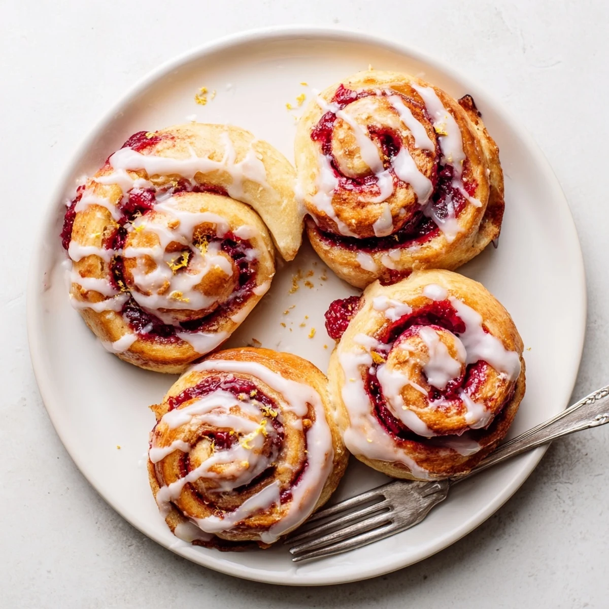 Golden-brown Raspberry Cinnamon Rolls With Lemon Glaze are drizzled with zesty icing on a white plate.
