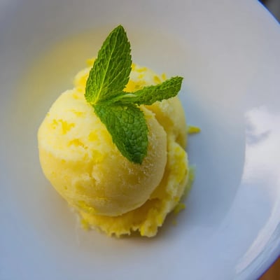A scoop of vibrant lemonade sorbet garnished with mint, served in a rustic bowl beside sliced lemons on a wooden table.